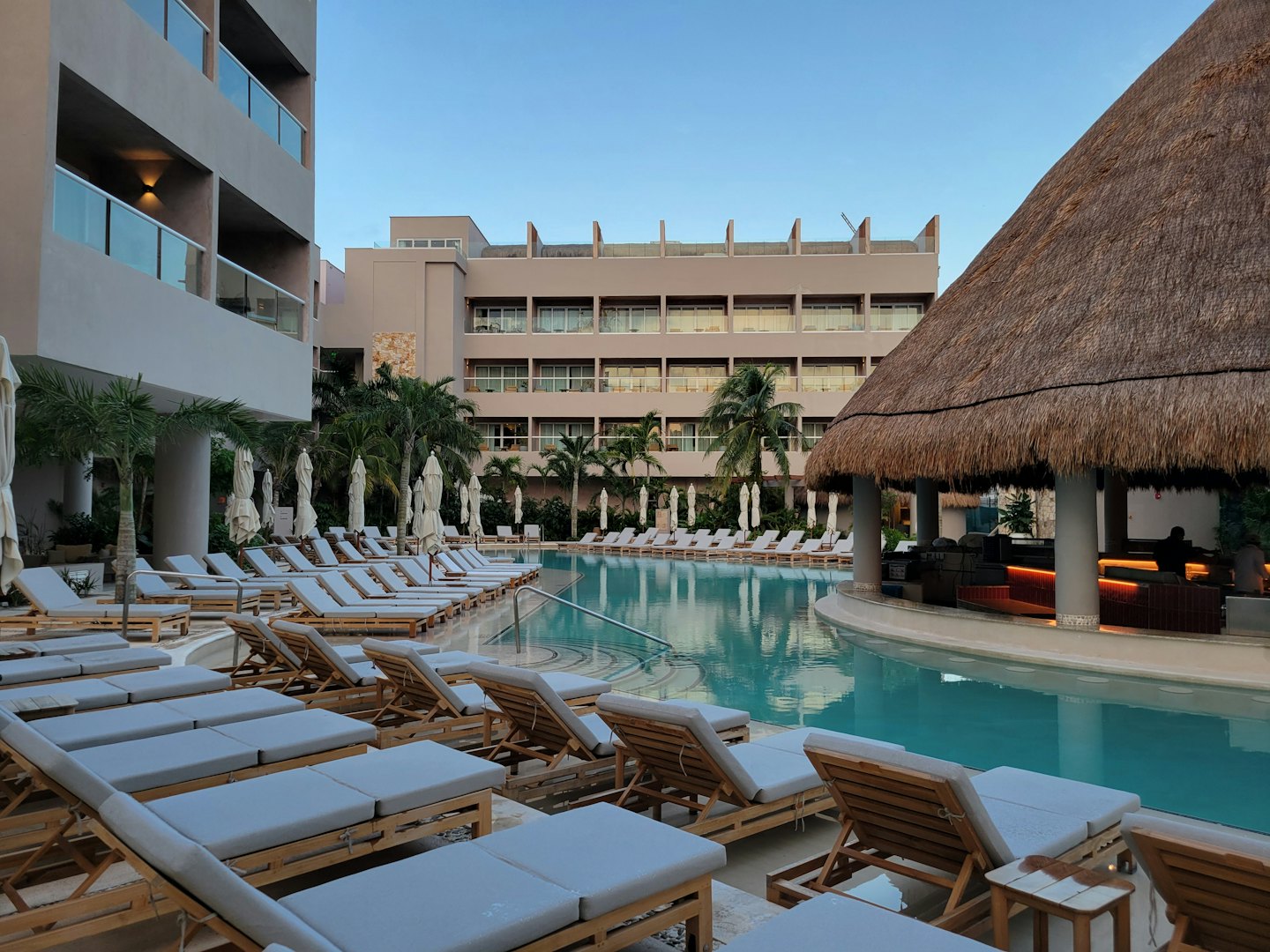 Resort swimming pool with empty lounge chairs and a thatched-roof bar.